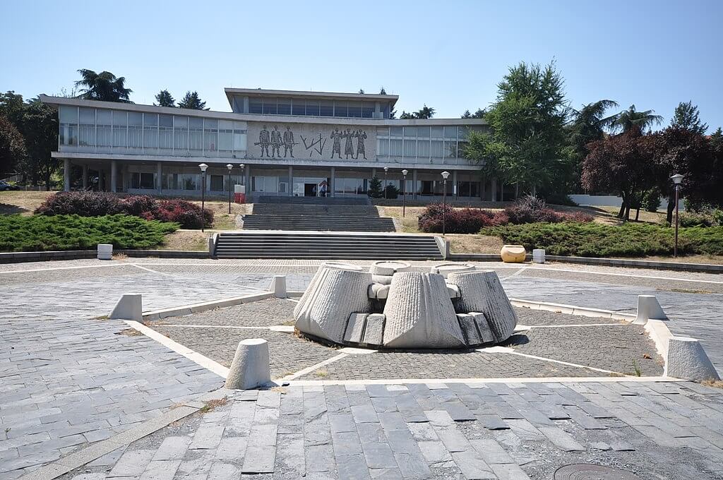 圖片中是位於塞爾維亞的南斯拉夫博物館，博物館正面設計簡約而大氣，前方有獨特的紀念雕塑與寬敞的廣場。